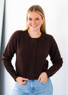 A smiling blonde woman wearing a dark brown button-front knit cardigan sweater and light blue jeans, standing against a plain light background.