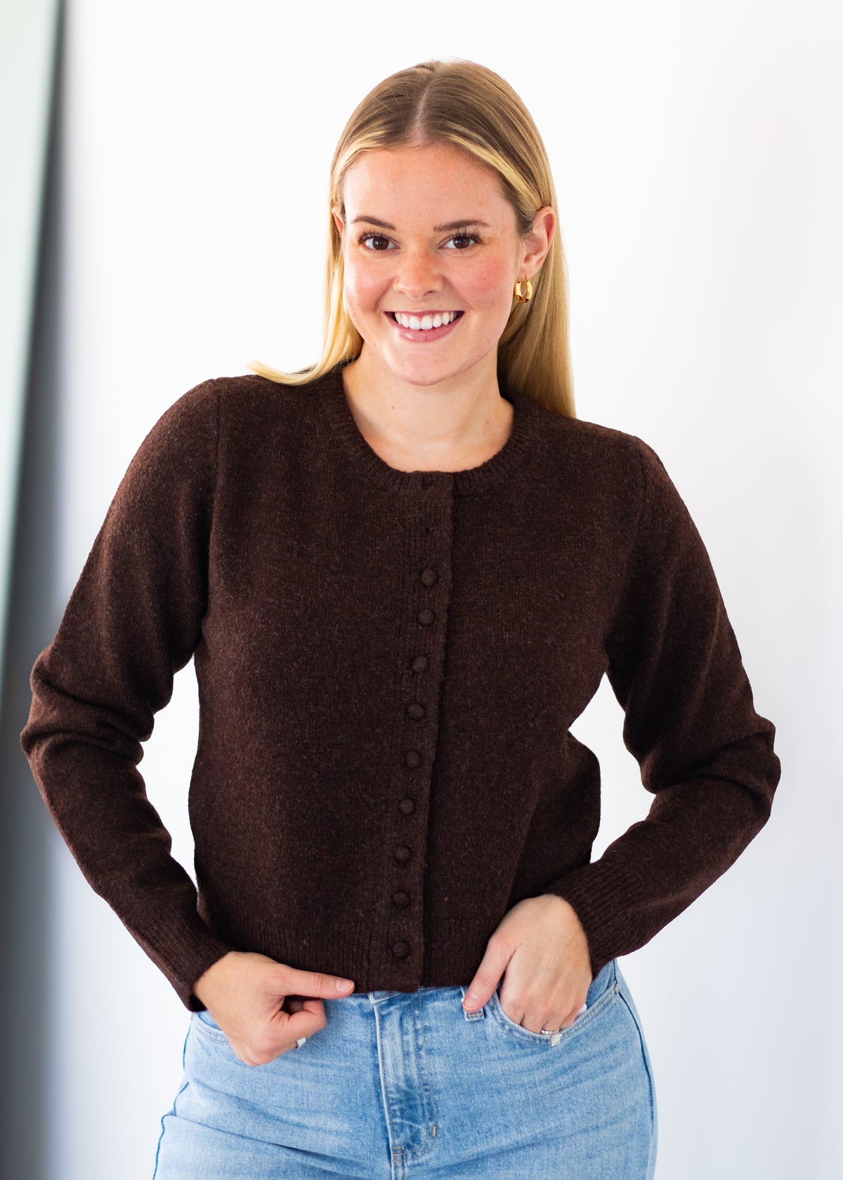 A smiling blonde woman wearing a dark brown button-front knit cardigan sweater and light blue jeans, standing against a plain light background.