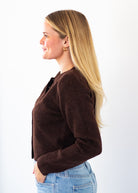 A woman with long blonde hair is shown in profile wearing a dark brown knit cardigan and light blue jeans against a plain white background.