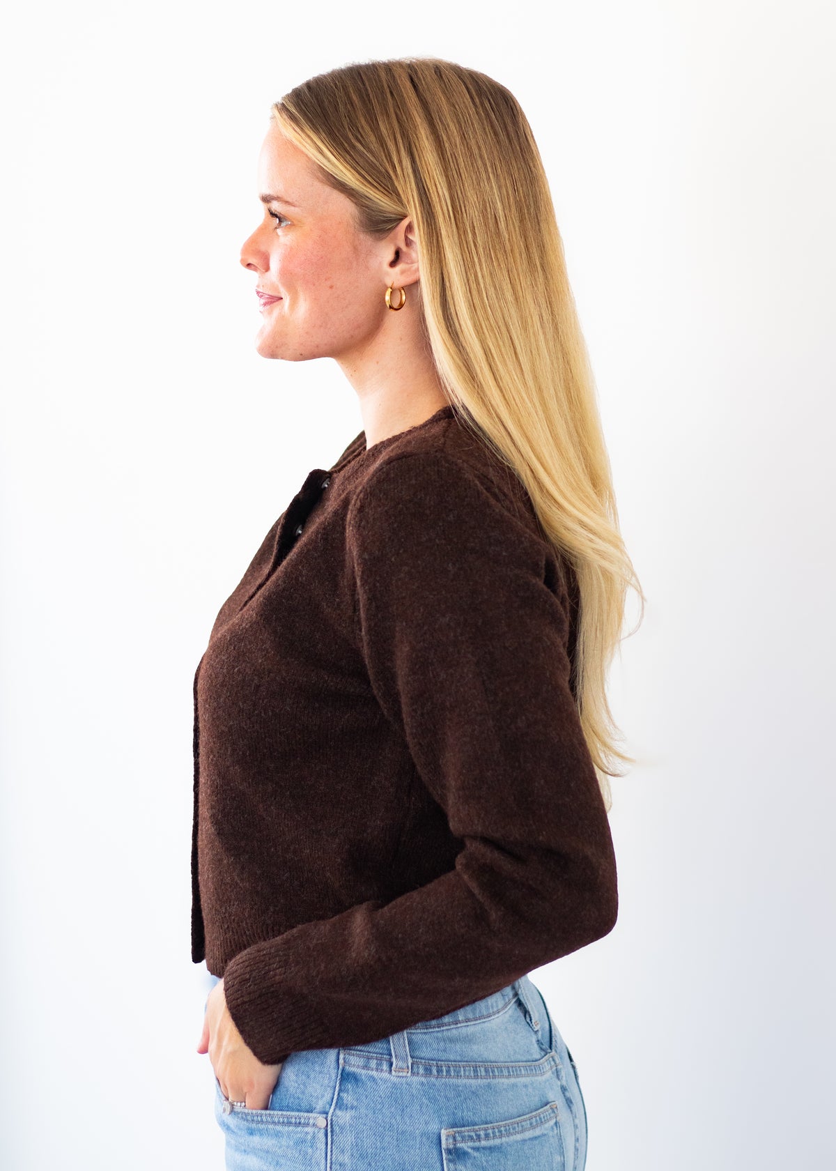 A woman with long blonde hair is shown in profile wearing a dark brown knit cardigan and light blue jeans against a plain white background.