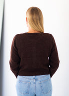 A woman viewed from behind wearing a dark brown knit sweater and light blue jeans against a plain white background.
