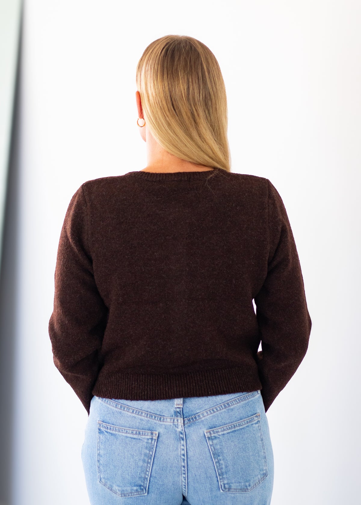 A woman viewed from behind wearing a dark brown knit sweater and light blue jeans against a plain white background.