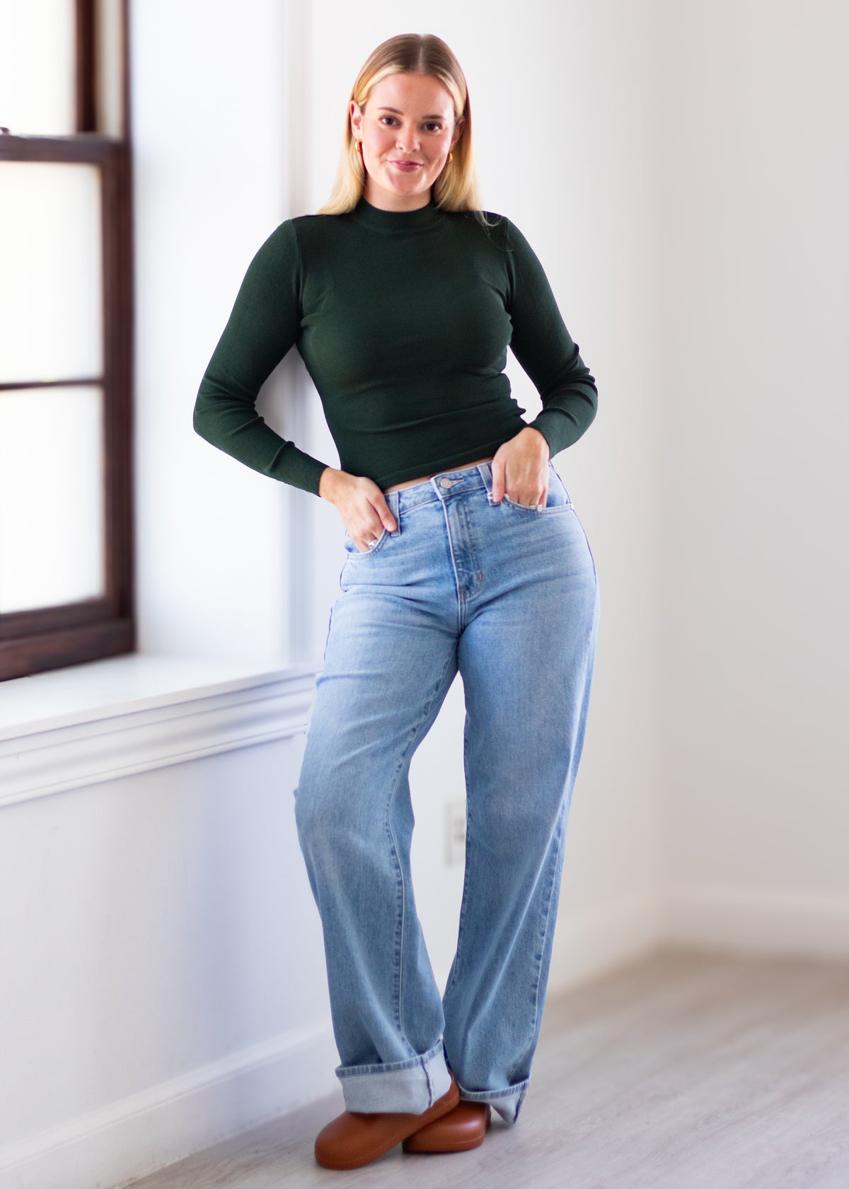 A woman modeling a dark green long-sleeve mock neck top and light blue wide-leg jeans, standing in a bright room with a window and light wood floor.