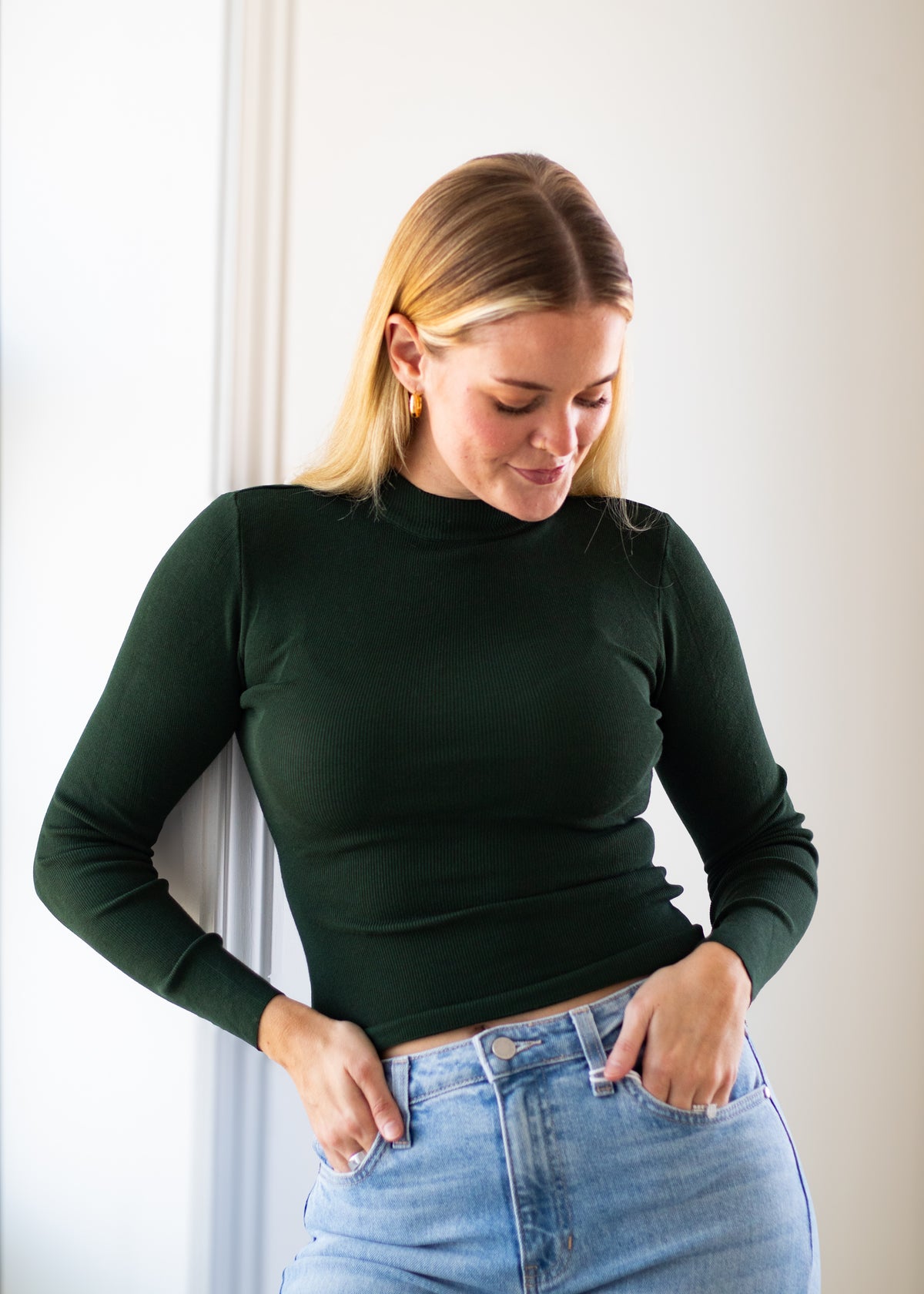 A woman wearing a dark forest green long-sleeve mock neck top and light wash blue jeans, standing against a white wall.