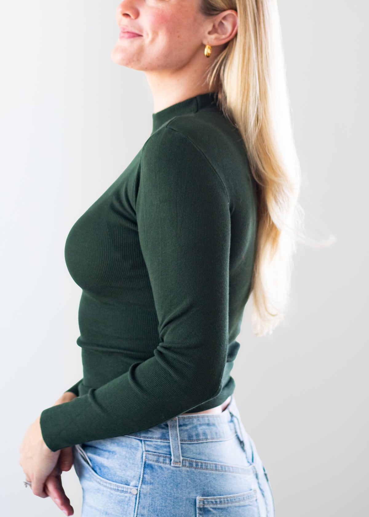 A woman shown in side profile wearing a fitted dark green mock-neck long-sleeve top and light blue jeans against a plain light gray background.