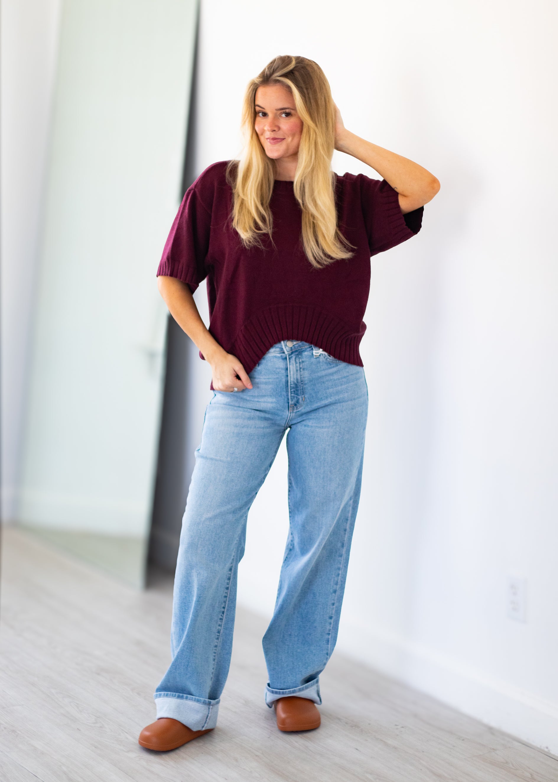 A woman modeling a short-sleeve burgundy knit sweater and light blue wide-leg jeans, standing in a bright indoor room with white walls and light wood flooring.