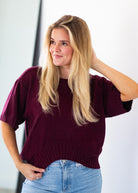 A woman with long blonde hair wearing a short-sleeve maroon knit sweater and light blue jeans, posing against a white background.