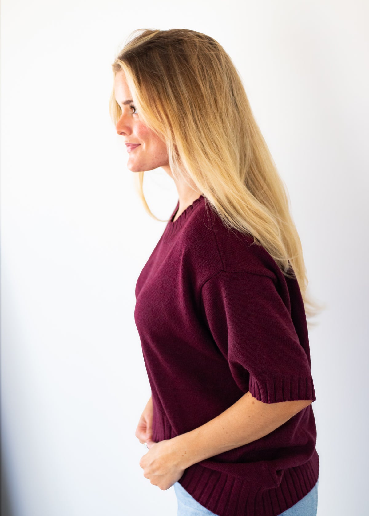A woman with long blonde hair is shown in profile wearing a short-sleeve knit sweater in deep burgundy against a plain white background.