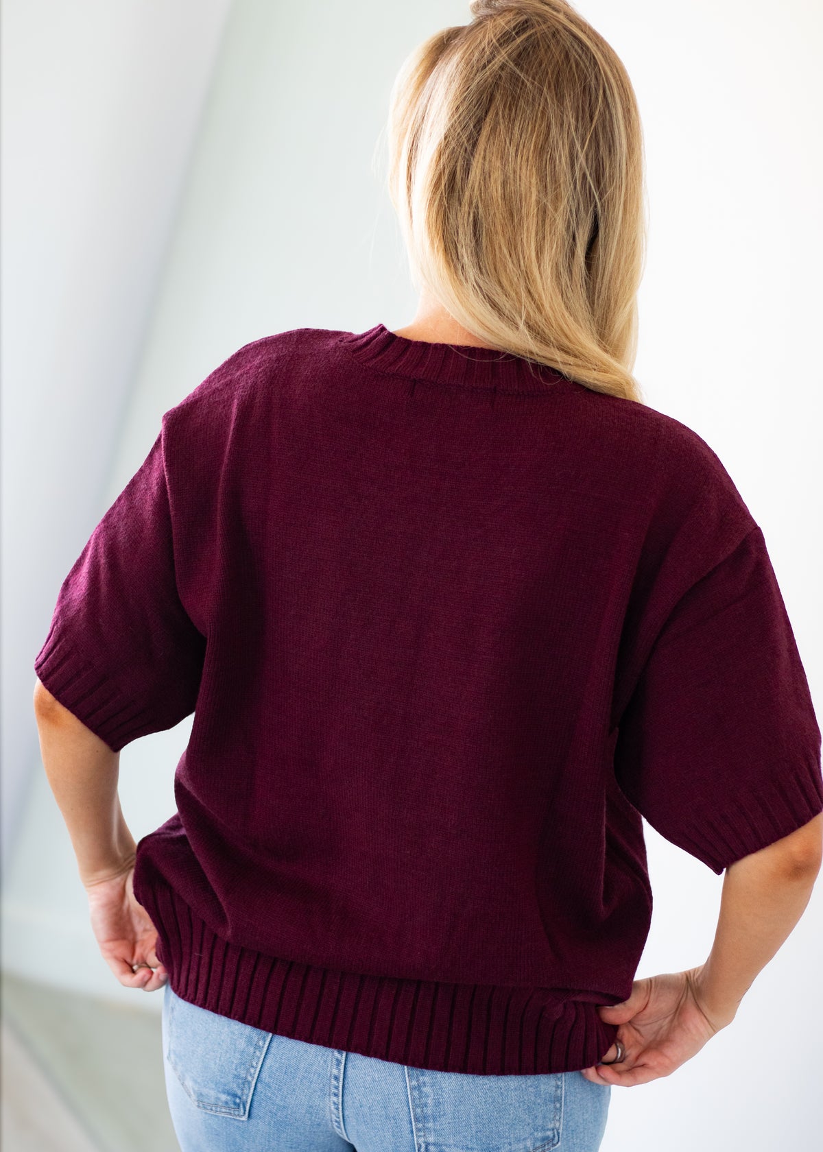 A woman viewed from behind wearing an oversized short-sleeve knit sweater in deep burgundy, paired with light blue jeans, against a clean white background.