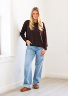 A woman with blonde hair stands in a bright room wearing a dark brown long-sleeve top, light blue wide-leg jeans, and brown leather shoes.