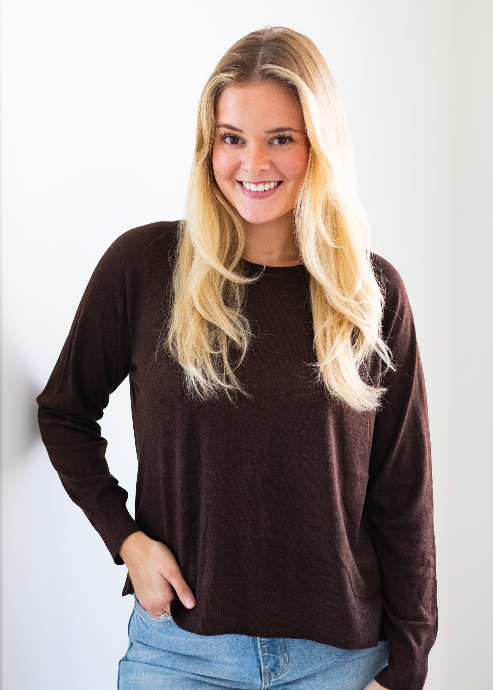 A smiling blonde woman wearing a dark brown long-sleeve crewneck top and light blue jeans, standing against a plain white background.