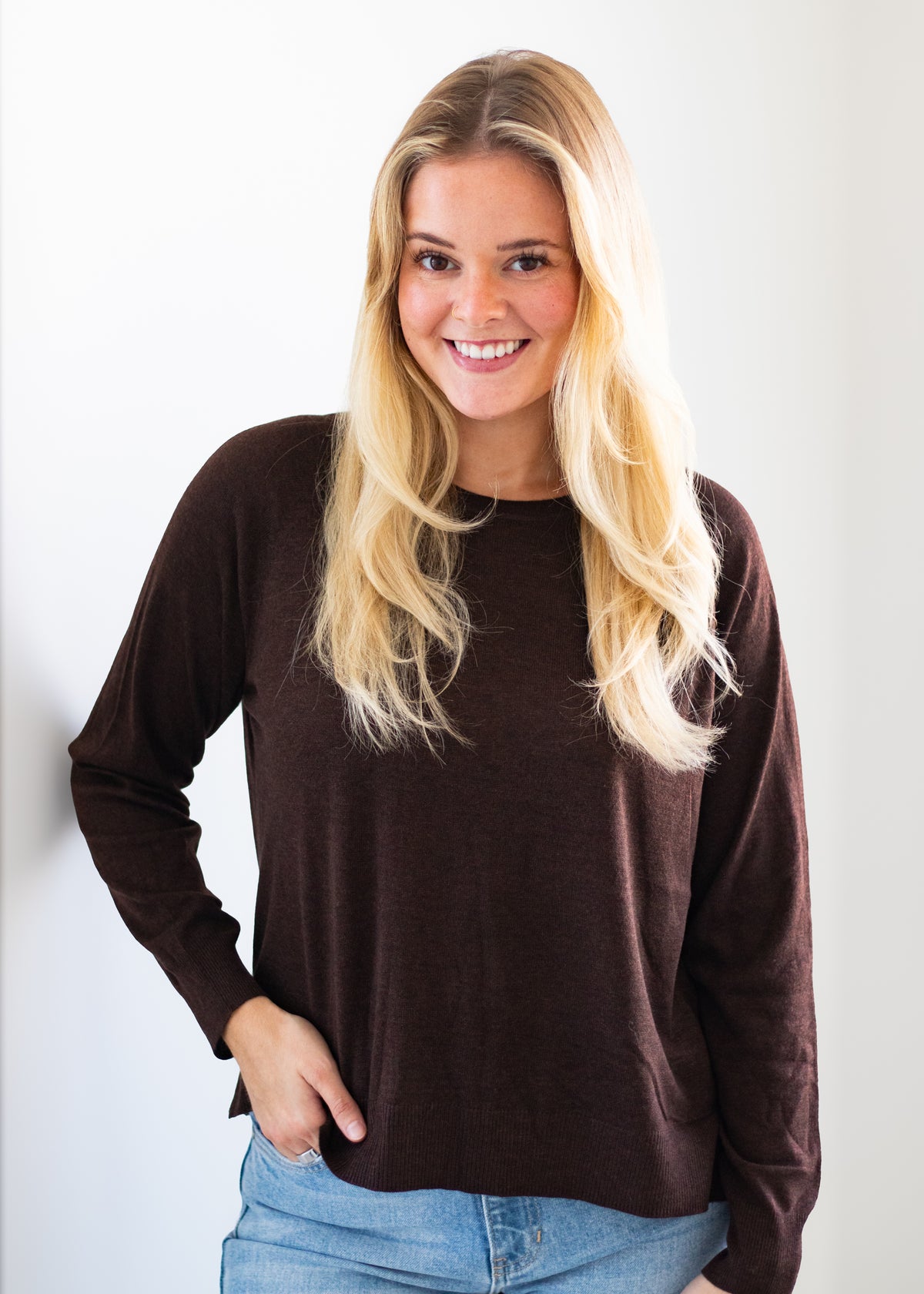 A smiling blonde woman wearing a dark brown long-sleeve crewneck top and light blue jeans, standing against a plain white background.