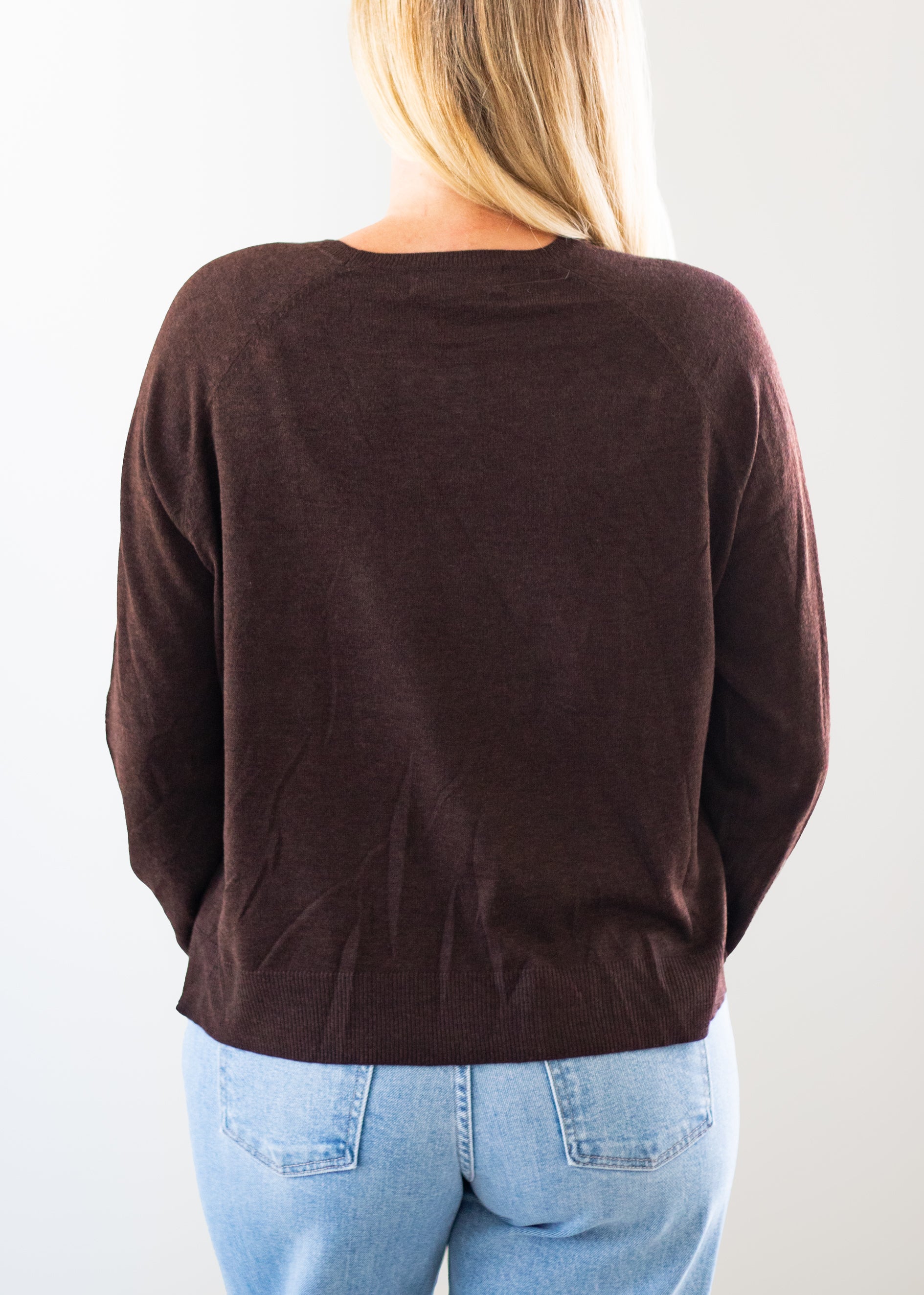 A woman viewed from behind wearing a dark brown long-sleeve knit top and light blue jeans against a plain white background.
