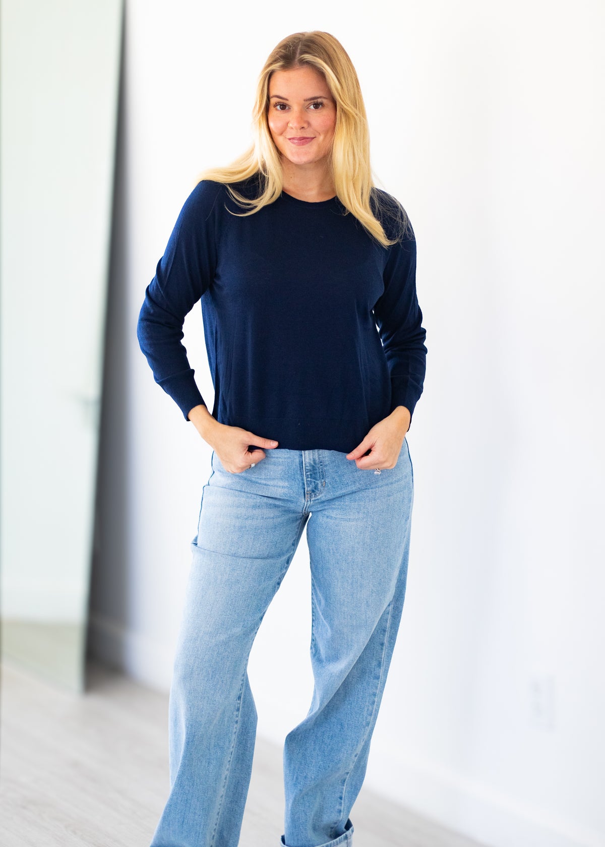 A woman modeling a navy blue long-sleeve crewneck top with light blue jeans, standing in a brightly lit indoor space with white walls.