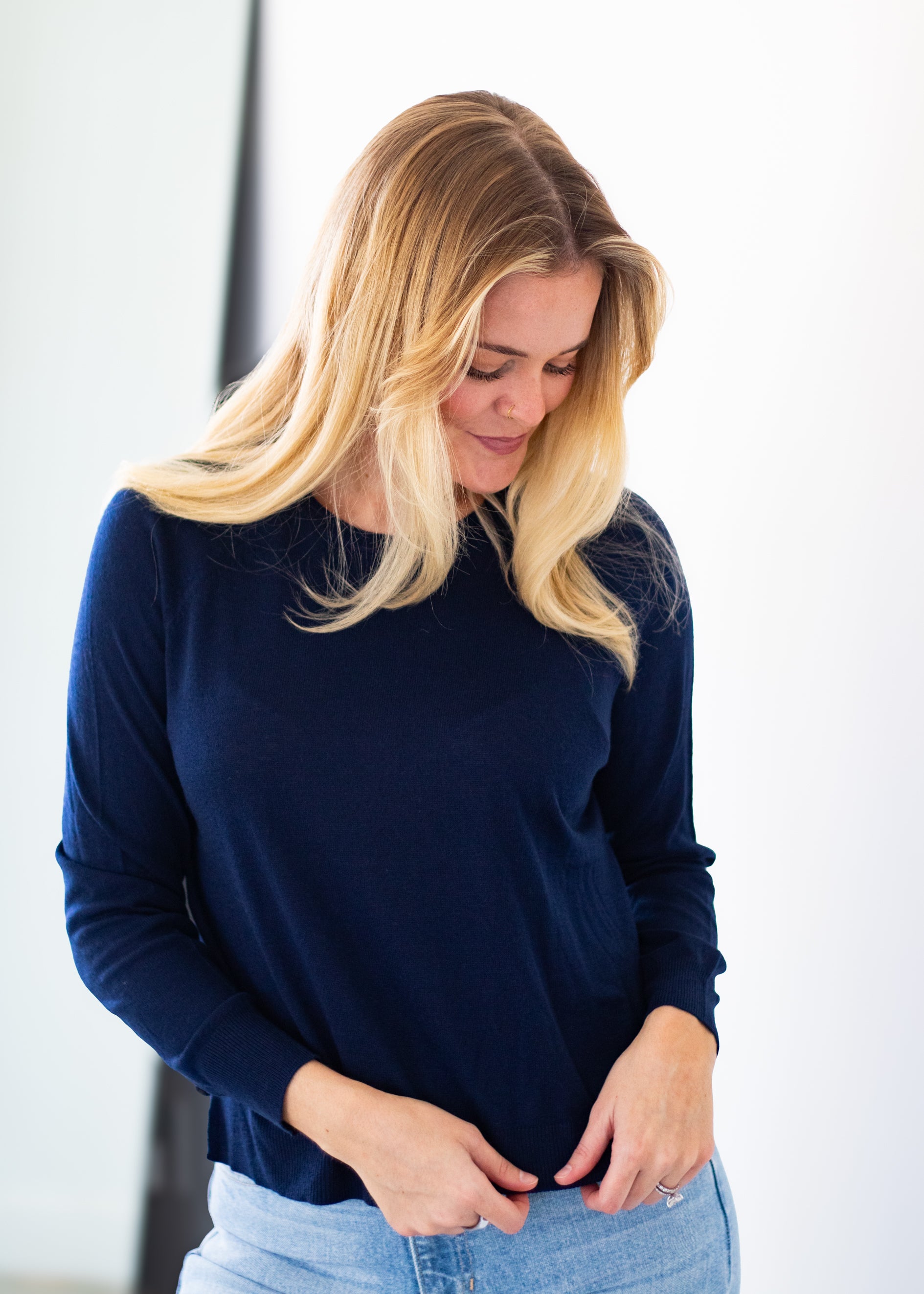 A blonde woman wearing a navy blue long-sleeve knit top and light blue jeans, looking down as she adjusts the front of her top.