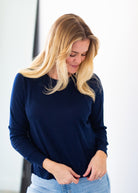 A blonde woman wearing a navy blue long-sleeve knit top and light blue jeans, looking down as she adjusts the front of her top.