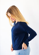 A woman with blonde hair wearing a navy blue long-sleeve knit top and light wash blue jeans, standing against a plain white background.