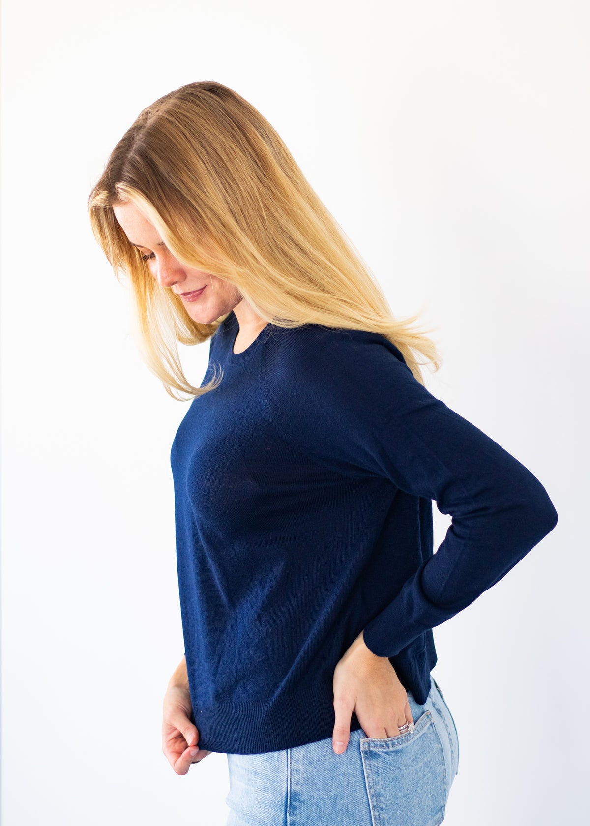 A woman with blonde hair wearing a navy blue long-sleeve knit top and light wash blue jeans, standing against a plain white background.