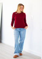 A woman wearing a long-sleeve crewneck knit sweater in deep burgundy, paired with light-wash blue jeans and brown leather shoes, standing in a bright, minimalist room with white walls and light wood flooring.