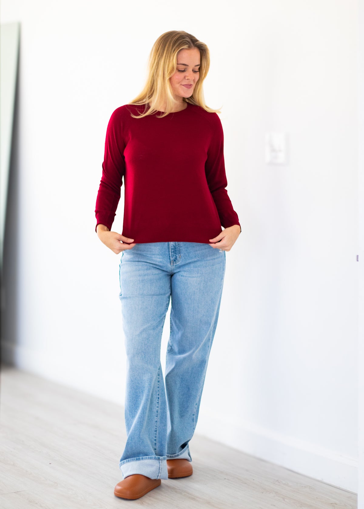 A woman wearing a long-sleeve crewneck knit sweater in deep burgundy, paired with light-wash blue jeans and brown leather shoes, standing in a bright, minimalist room with white walls and light wood flooring.