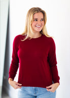 A smiling blonde woman wearing a long-sleeve crewneck knit sweater in deep burgundy, paired with light blue jeans, standing against a bright white background.