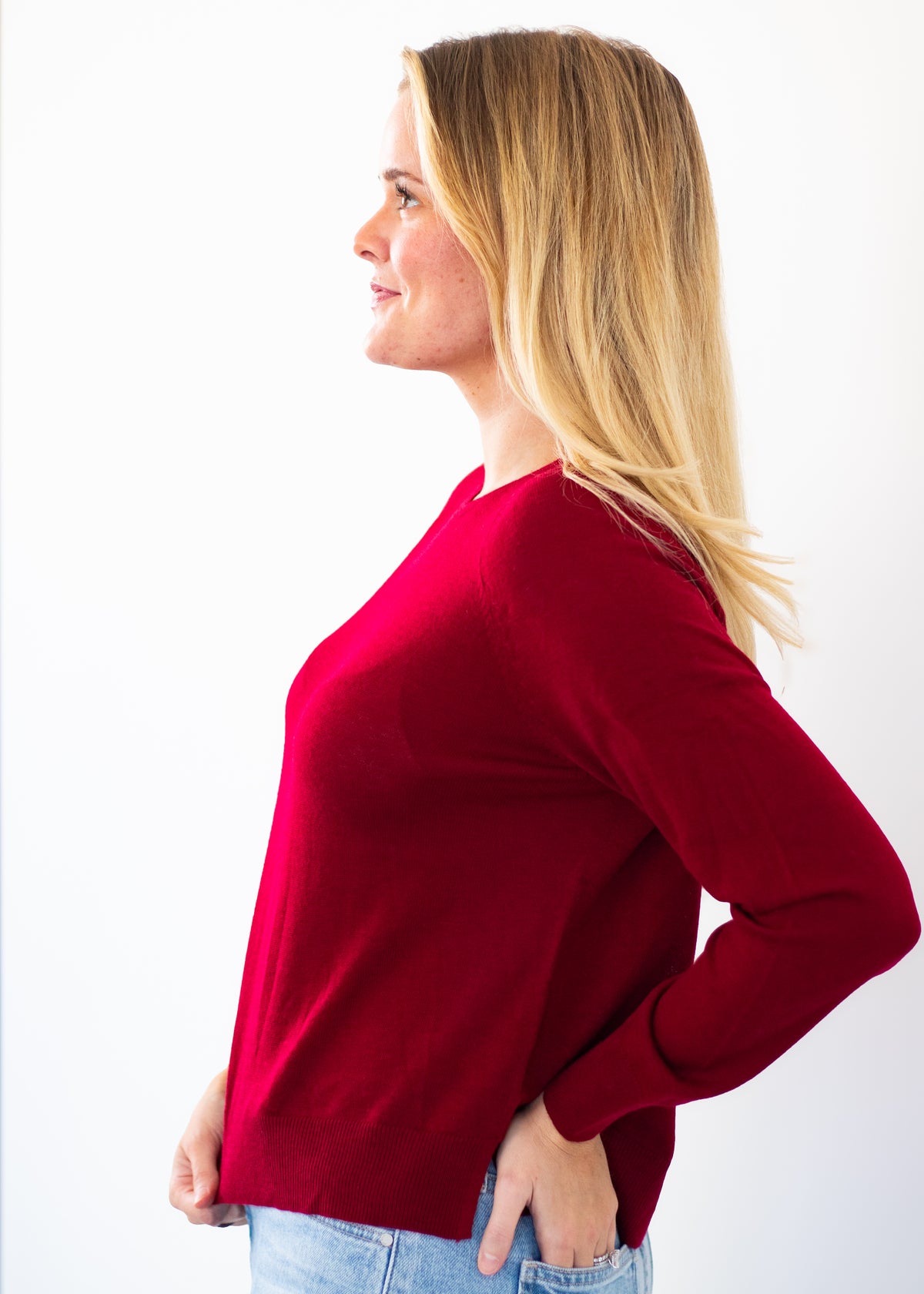 A woman with blonde hair is shown in profile wearing a long-sleeve knit top in a deep red or burgundy color, paired with light blue jeans, against a plain white background.