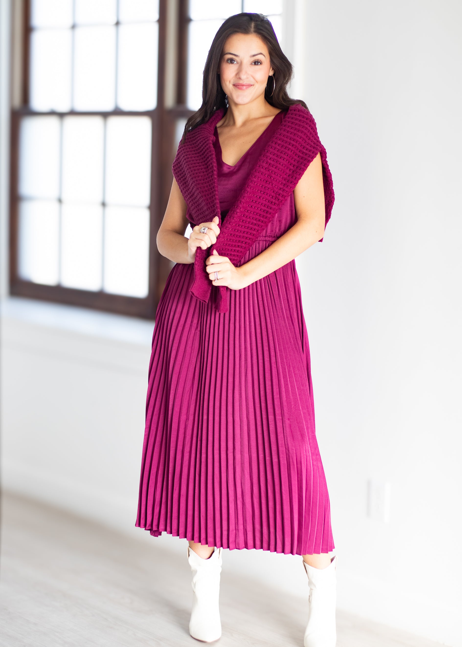 A woman modeling a bright magenta pleated midi dress with a matching knit shawl draped over her shoulders, paired with white ankle boots, in a bright room with a large window.