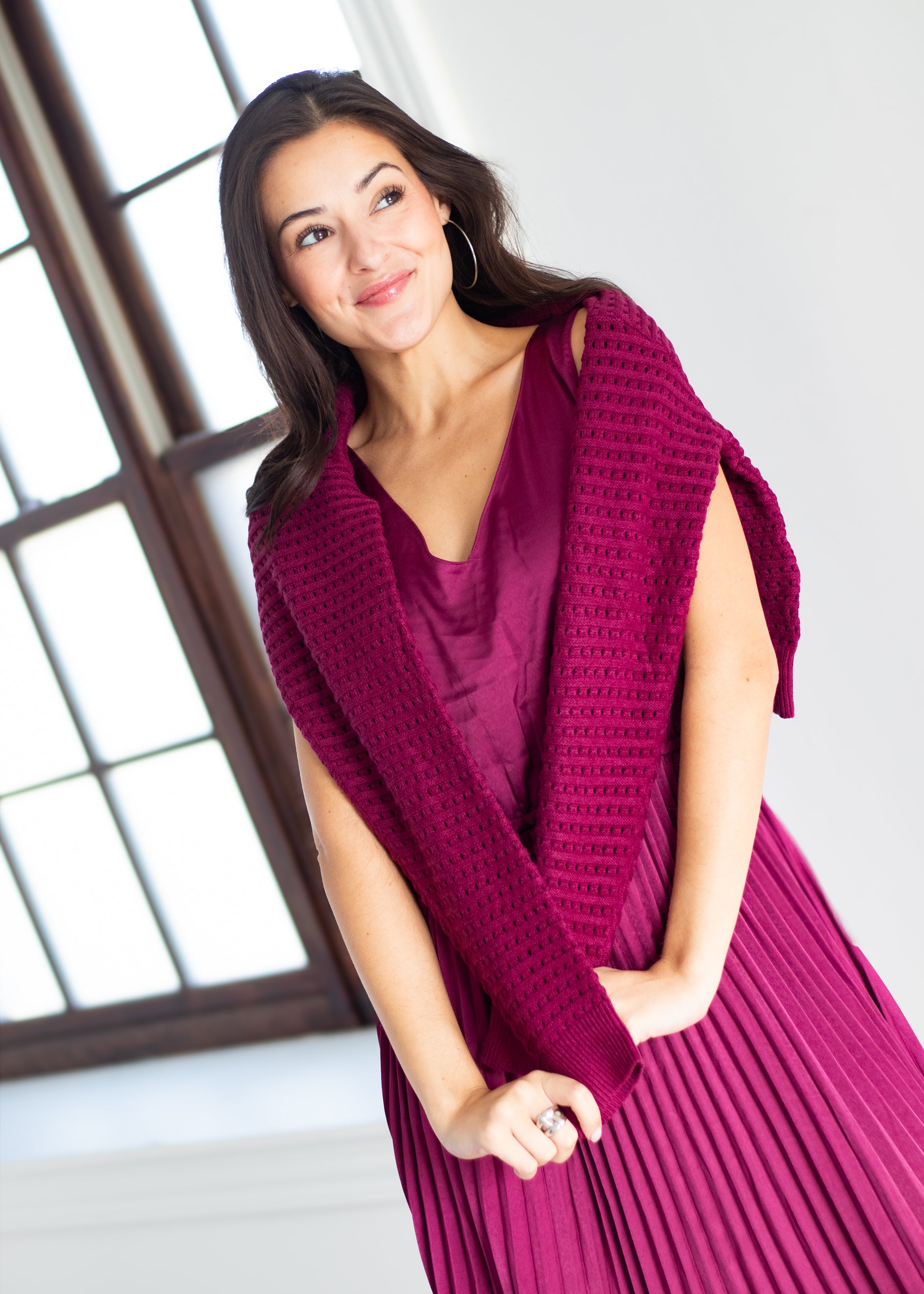 A woman wearing a deep magenta pleated dress with a matching open-knit cardigan draped over her shoulders, standing in a bright room with a window.