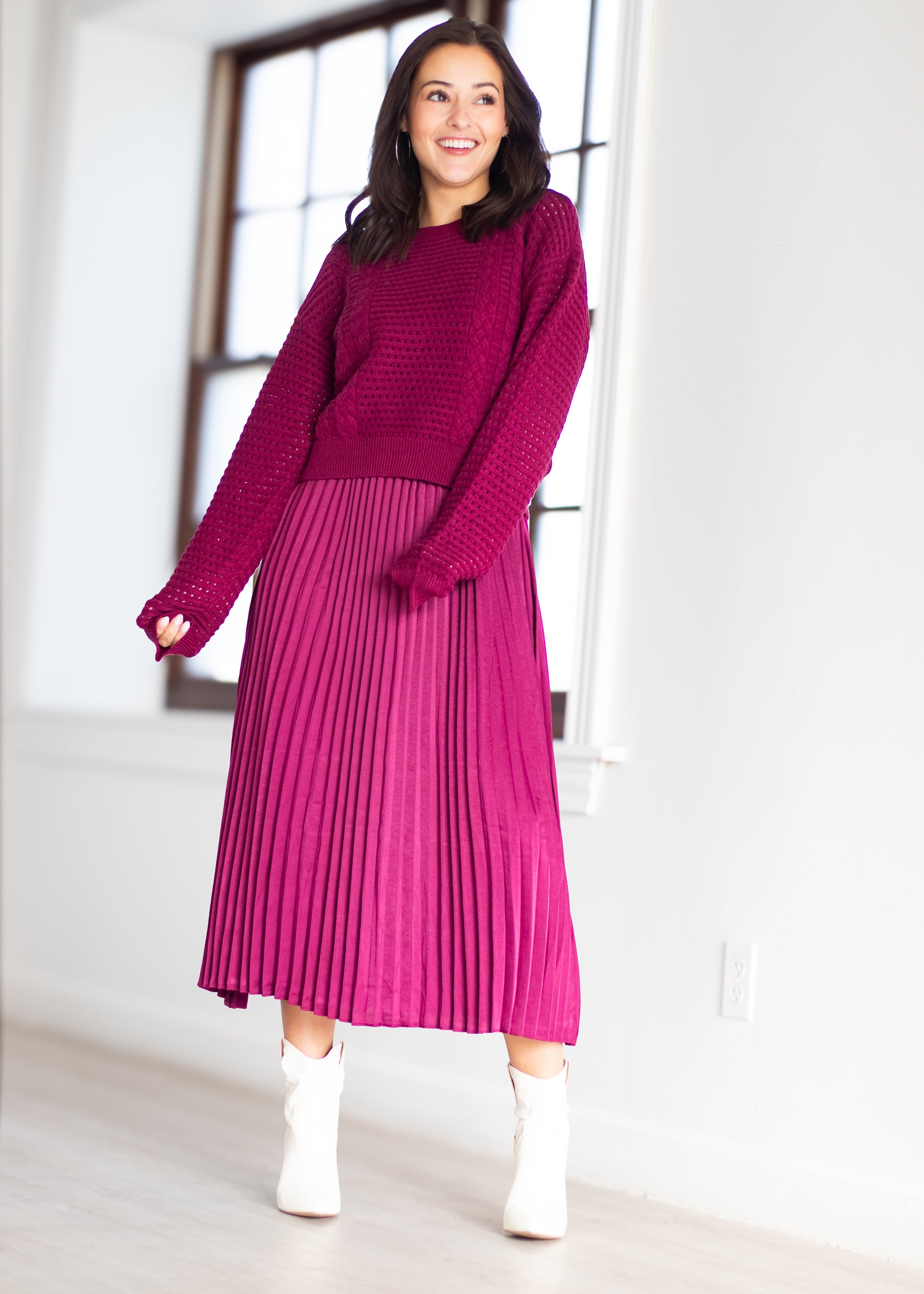 A woman modeling a two-piece knit set consisting of a textured magenta long-sleeve top and a matching pleated midi skirt, paired with white ankle boots in a bright room.