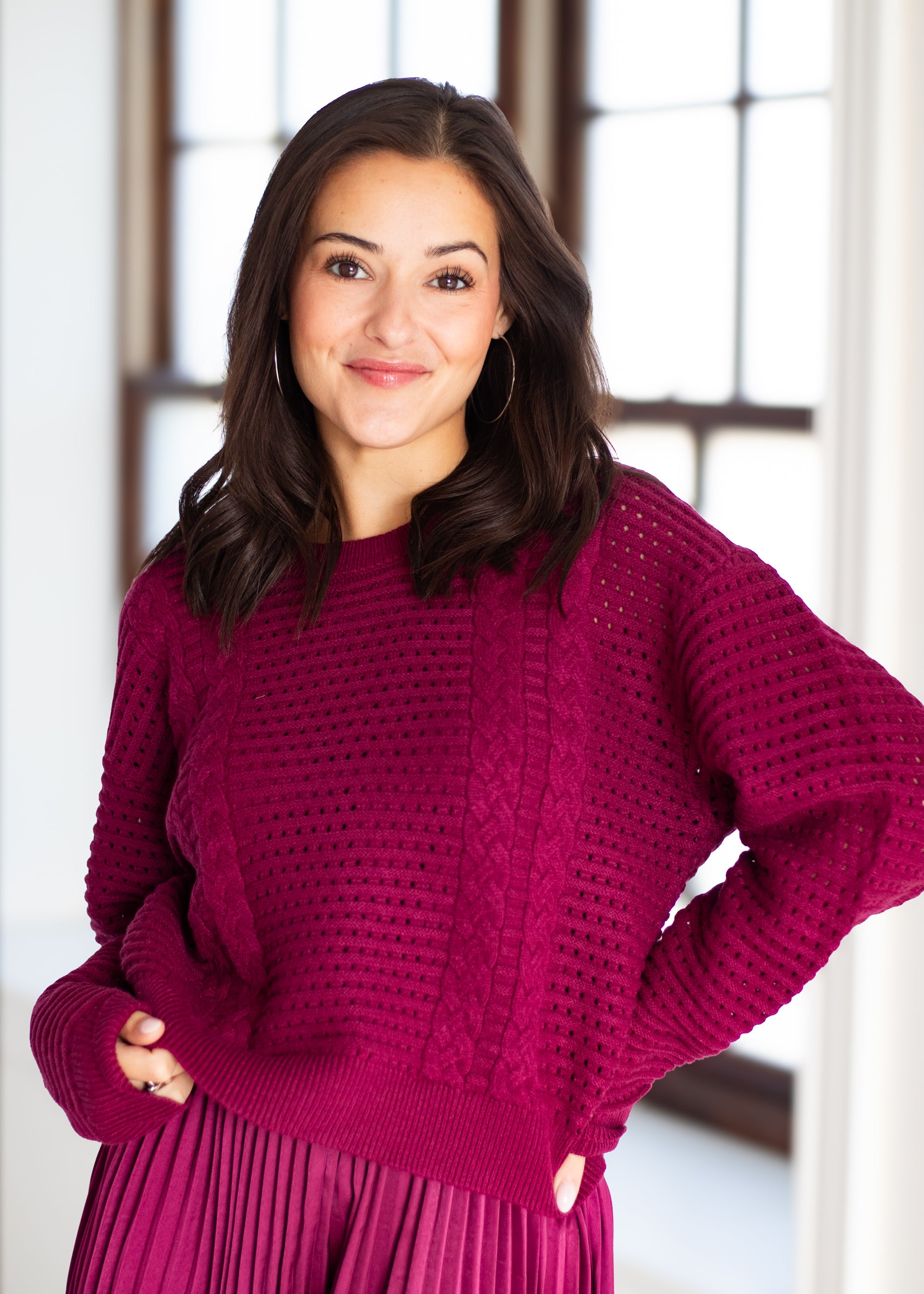 A woman wearing a chunky knit crewneck sweater in a rich magenta-purple color, paired with a matching pleated skirt, standing in a brightly lit room with large windows.