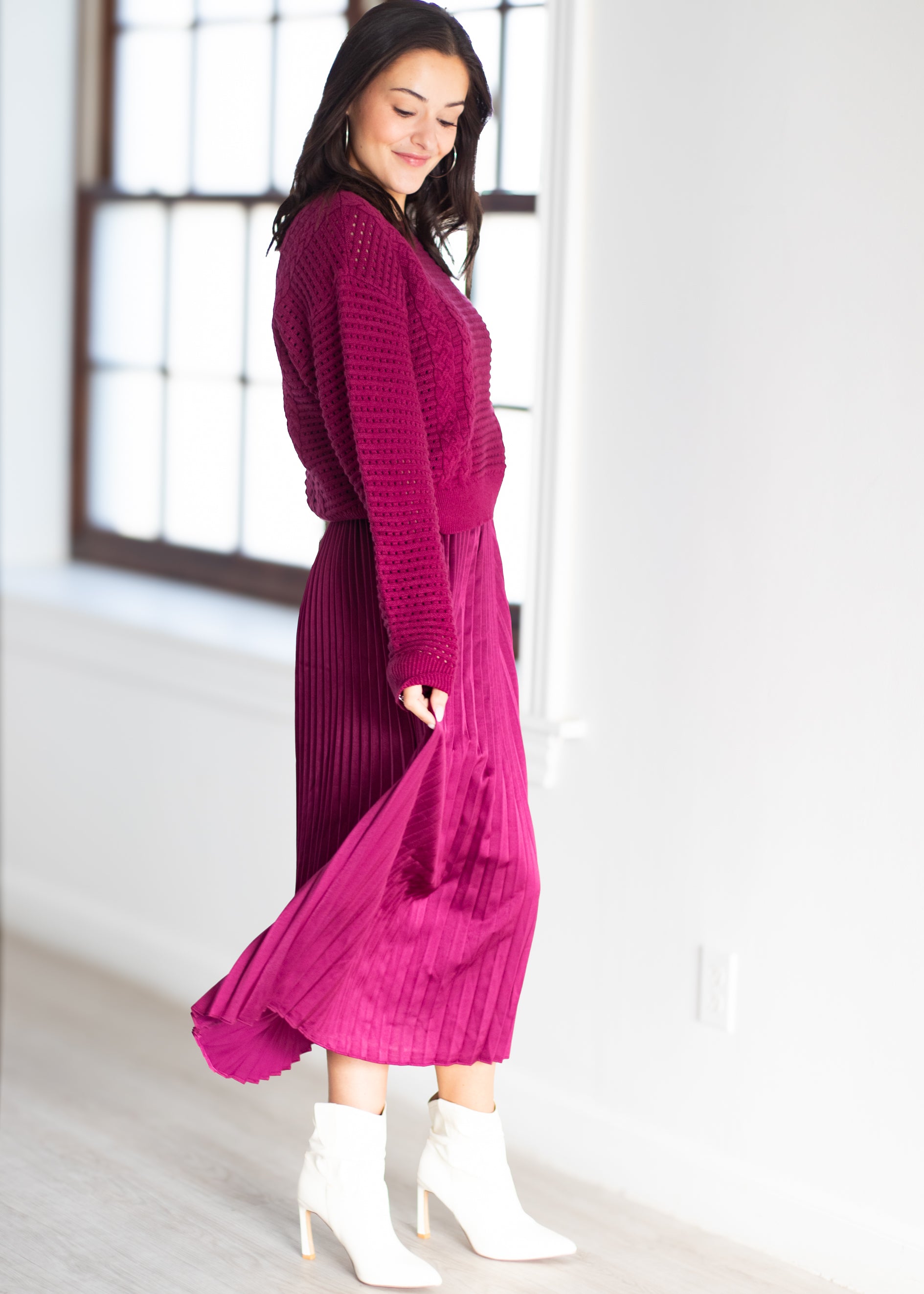 A woman wearing a vibrant magenta two-piece knit and pleated dress with white ankle boots, standing in a bright room with a window.
