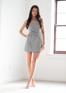 A woman modeling a black and white gingham strapless mini dress with a tie waist, standing in a bright white room with a window.