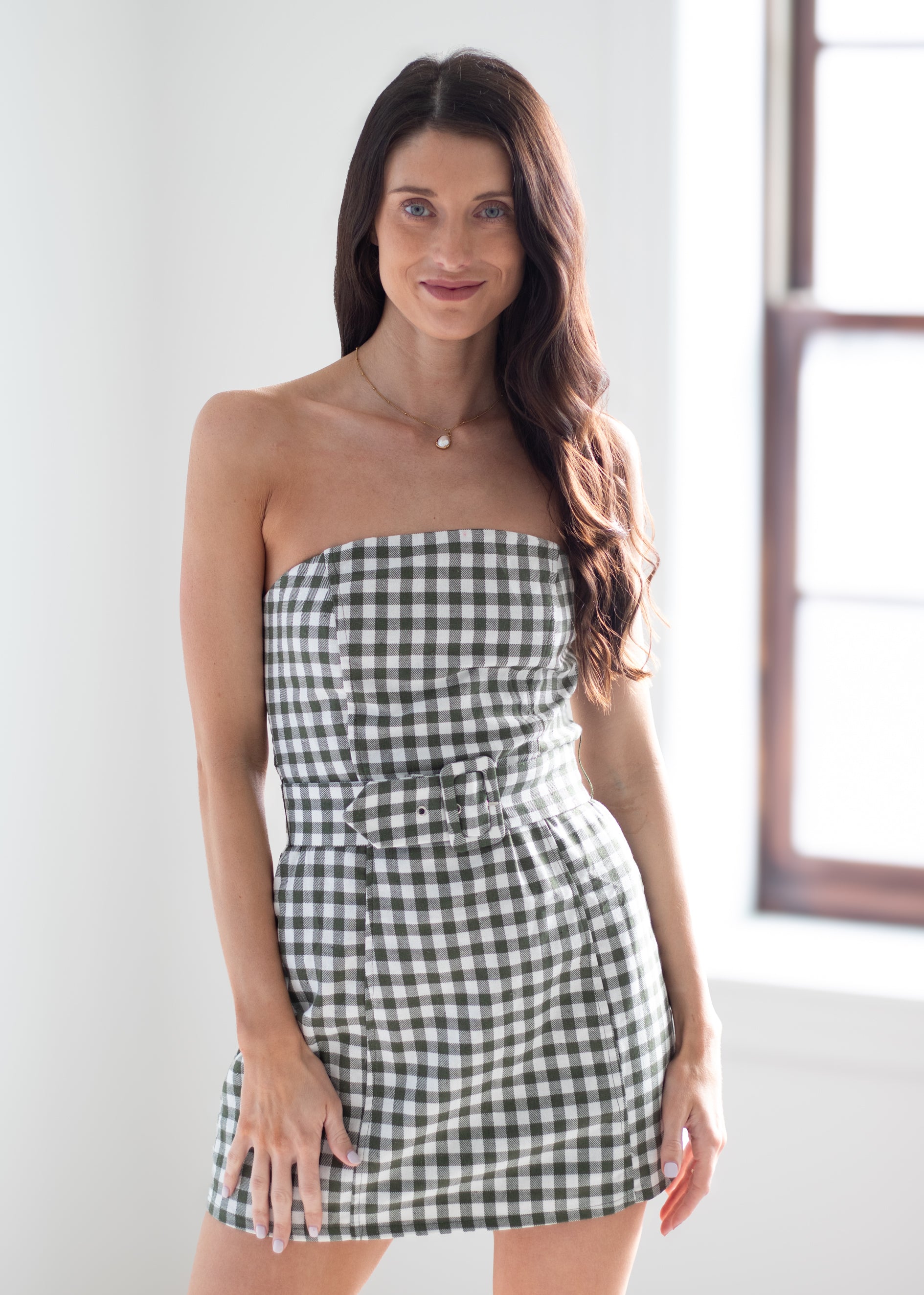 A woman modeling a strapless black and white gingham mini dress with a matching fabric belt, standing in a brightly lit room with a window in the background.