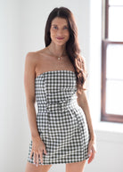 A woman modeling a strapless black and white gingham mini dress with a matching fabric belt, standing in a brightly lit room with a window in the background.