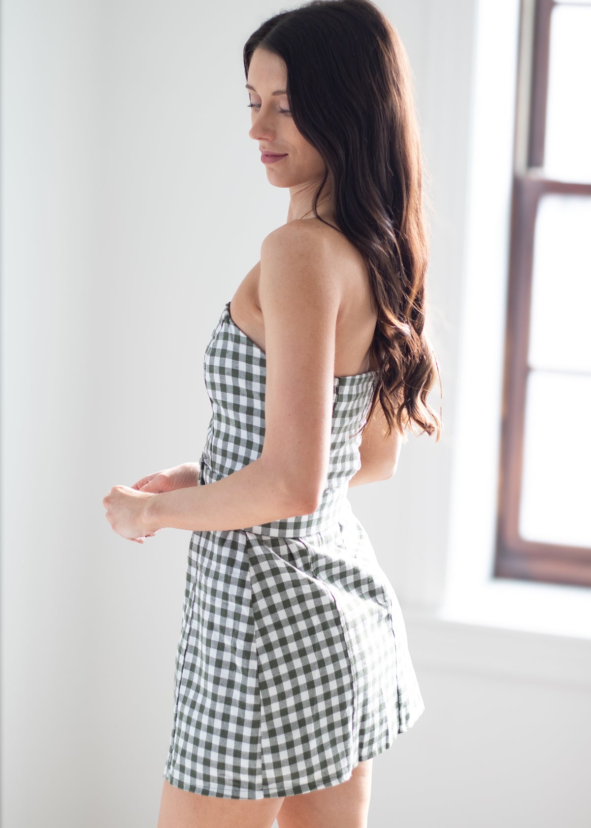 A woman wearing a black and white gingham strapless mini dress, standing in a bright room with a window on the right.