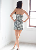 A woman stands with her back to the camera wearing a strapless black and white gingham mini dress in a bright, white room with a window.