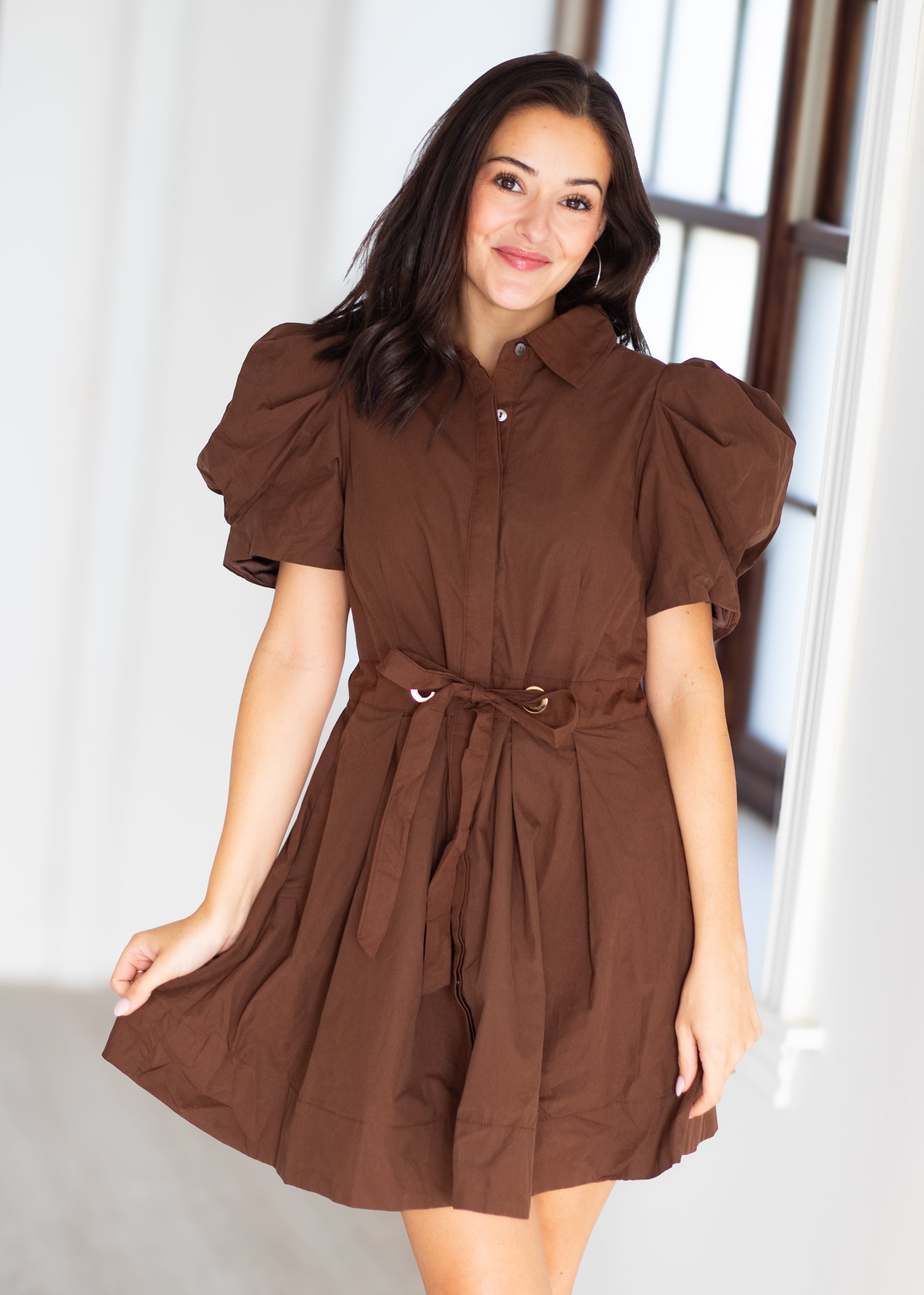A woman wearing a brown short puff-sleeve dress with a button-down front, collar, and matching tie belt at the waist, standing in a bright indoor setting.