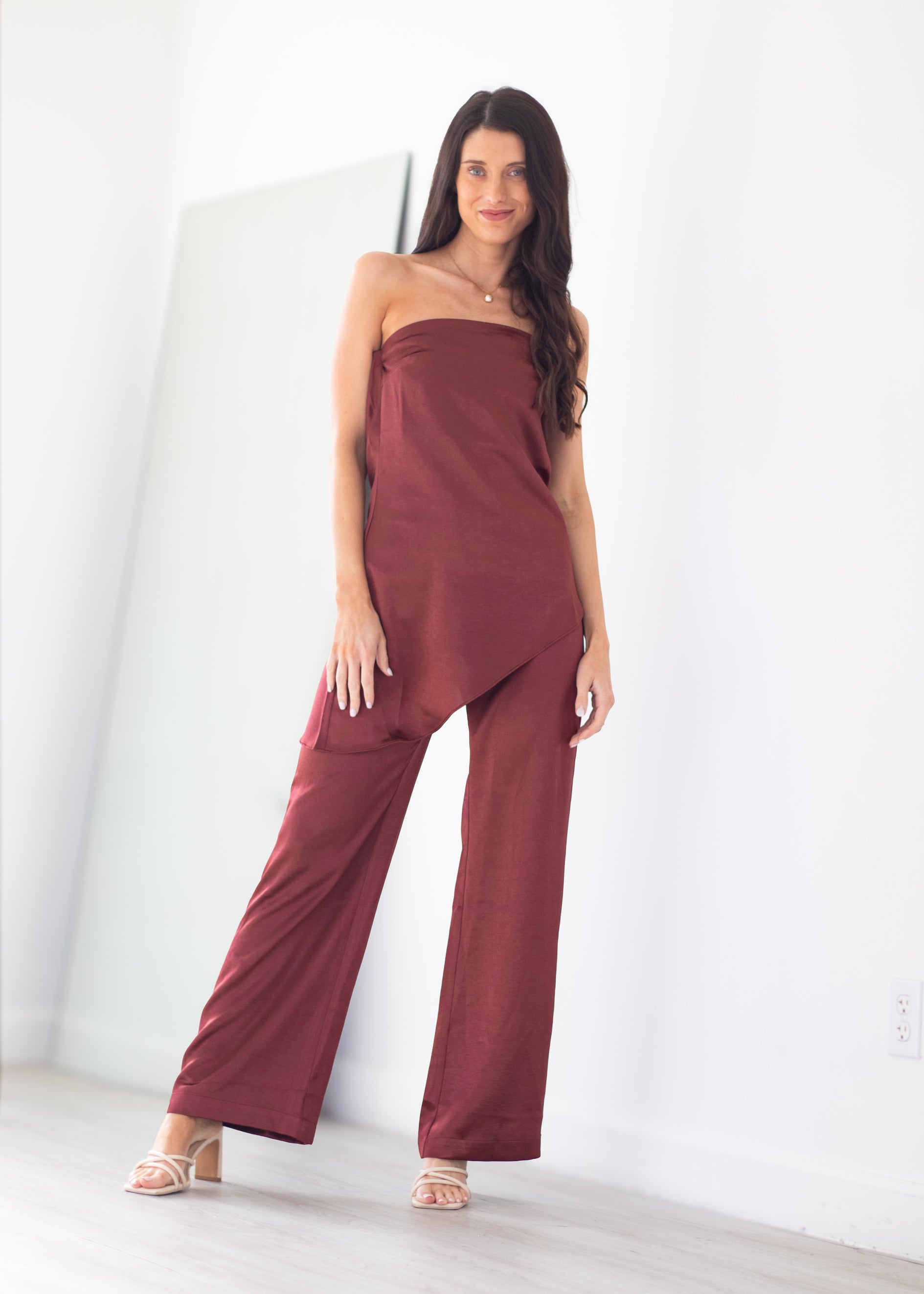 A woman modeling a strapless wide-leg jumpsuit in a solid deep burgundy color, standing in a brightly lit minimalist room with white walls and light wood flooring.
