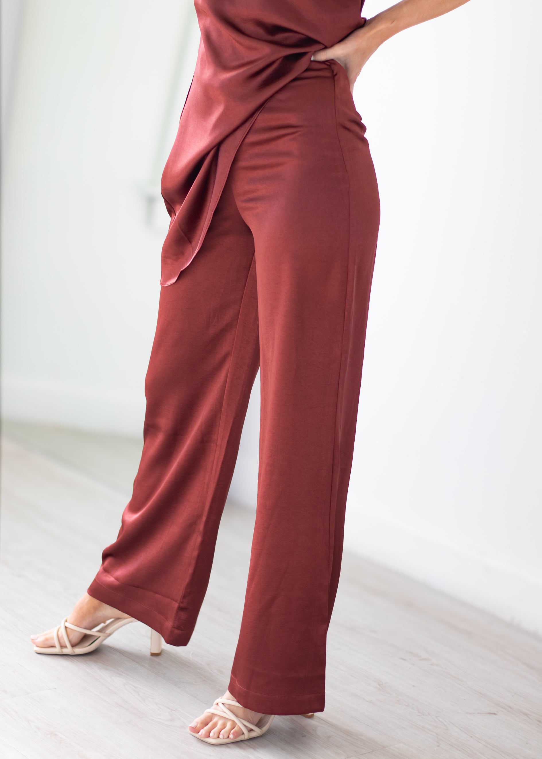 A person wearing high-waisted, wide-leg trousers in a dusty rose or muted terracotta color, paired with a matching draped top and light-colored heeled sandals, standing in a minimalist indoor setting.