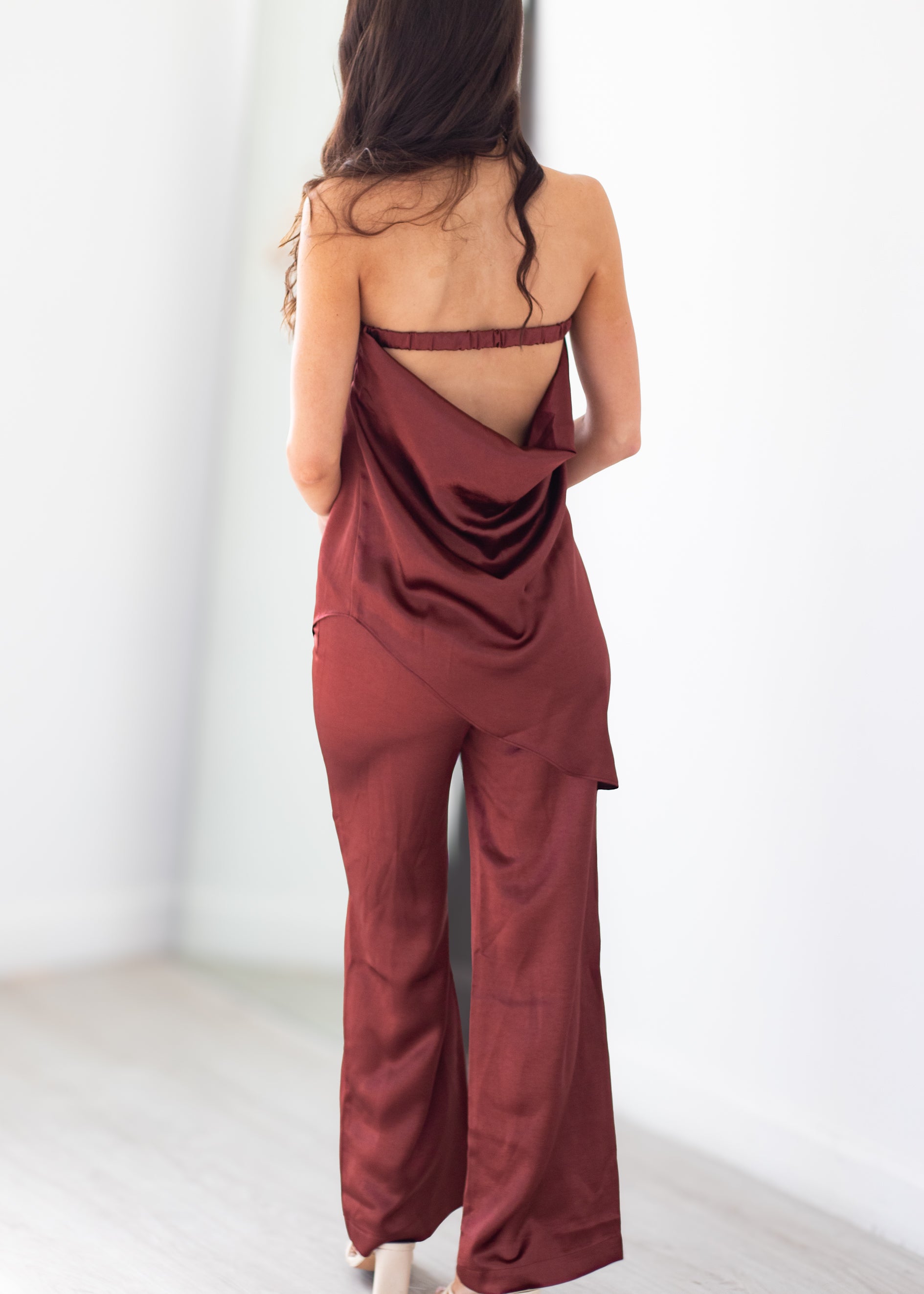 A woman shown from behind wearing a rust-brown satin-like jumpsuit with a draped open back and wide legs, standing in a bright white indoor space.