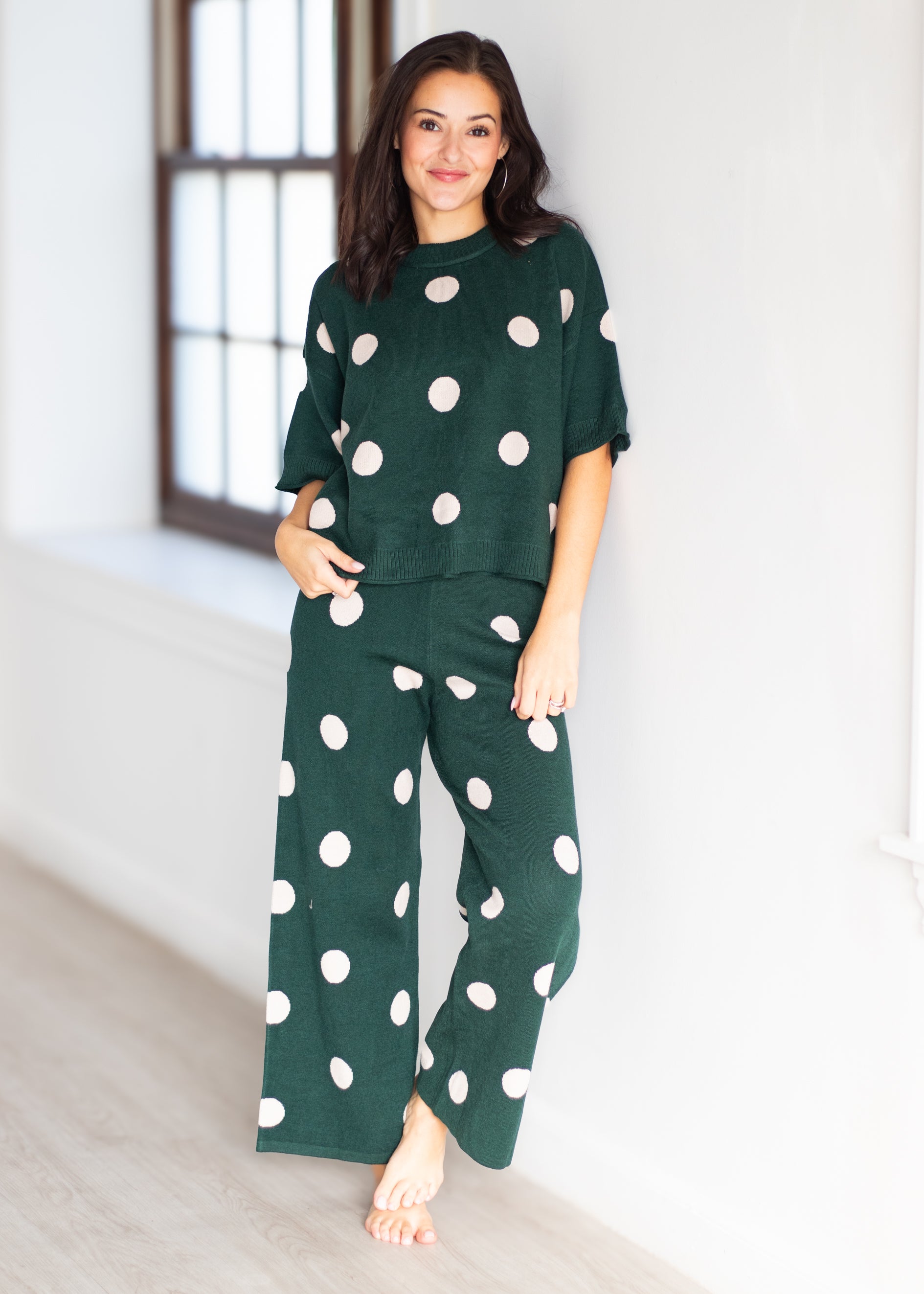 A woman modeling a dark green two-piece pajama set with large white polka dots, consisting of a short-sleeve top and matching wide-leg pants, standing in a bright room.