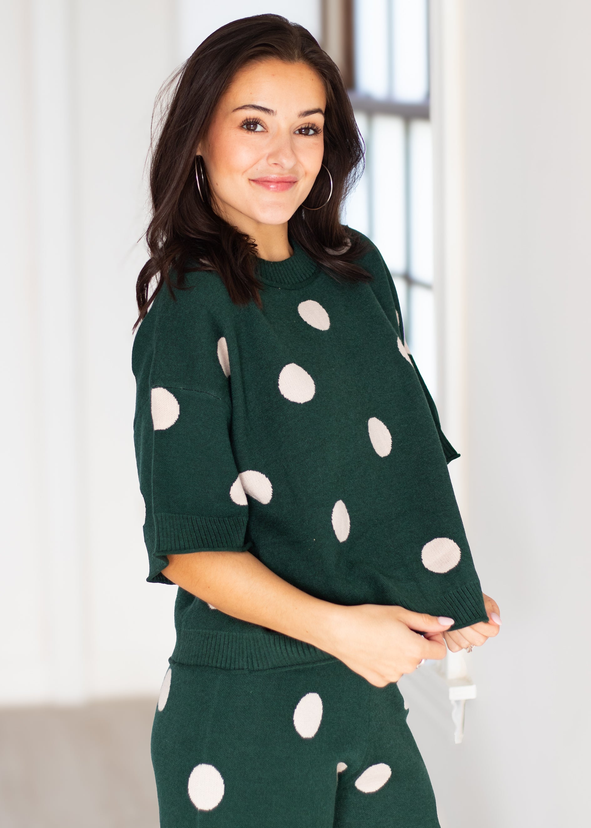 A woman wearing a dark green knit sweater and matching pants with large white polka dots, standing in a bright indoor setting.