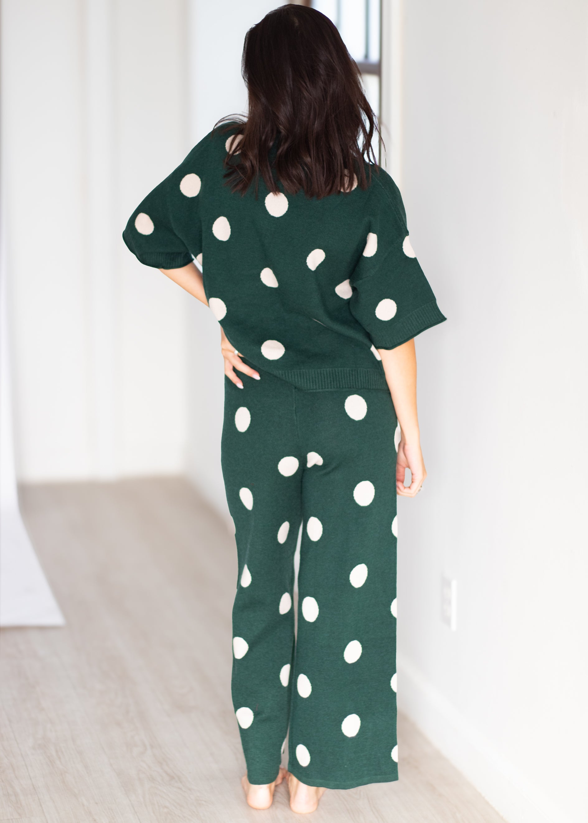A woman viewed from behind wearing a dark green two-piece lounge set with large white polka dots, consisting of a short-sleeve top and matching wide-leg pants, in a brightly lit room with light wood flooring.