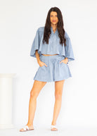 A woman modeling a light blue denim two-piece set consisting of a cropped long-sleeve blouse with wide bell sleeves and matching high-waisted shorts with a drawstring waist, set against a plain white studio background.