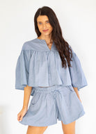 A woman modeling a light blue and white vertically striped two-piece set consisting of a short-sleeve cropped blouse with ruffles and matching drawstring shorts, set against a plain white background.