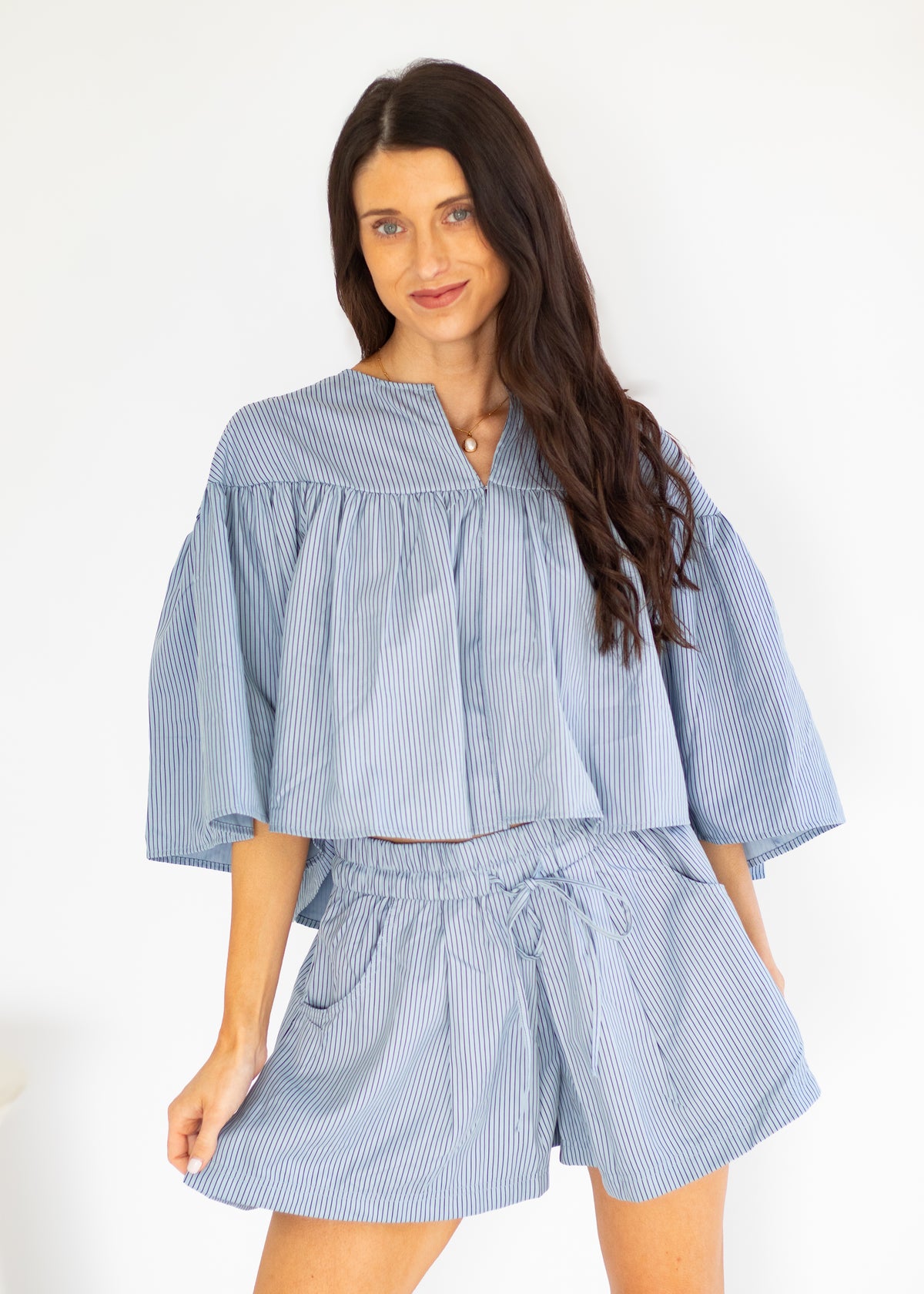 A woman modeling a light blue and white vertically striped two-piece set consisting of a short-sleeve cropped blouse with ruffles and matching drawstring shorts, set against a plain white background.