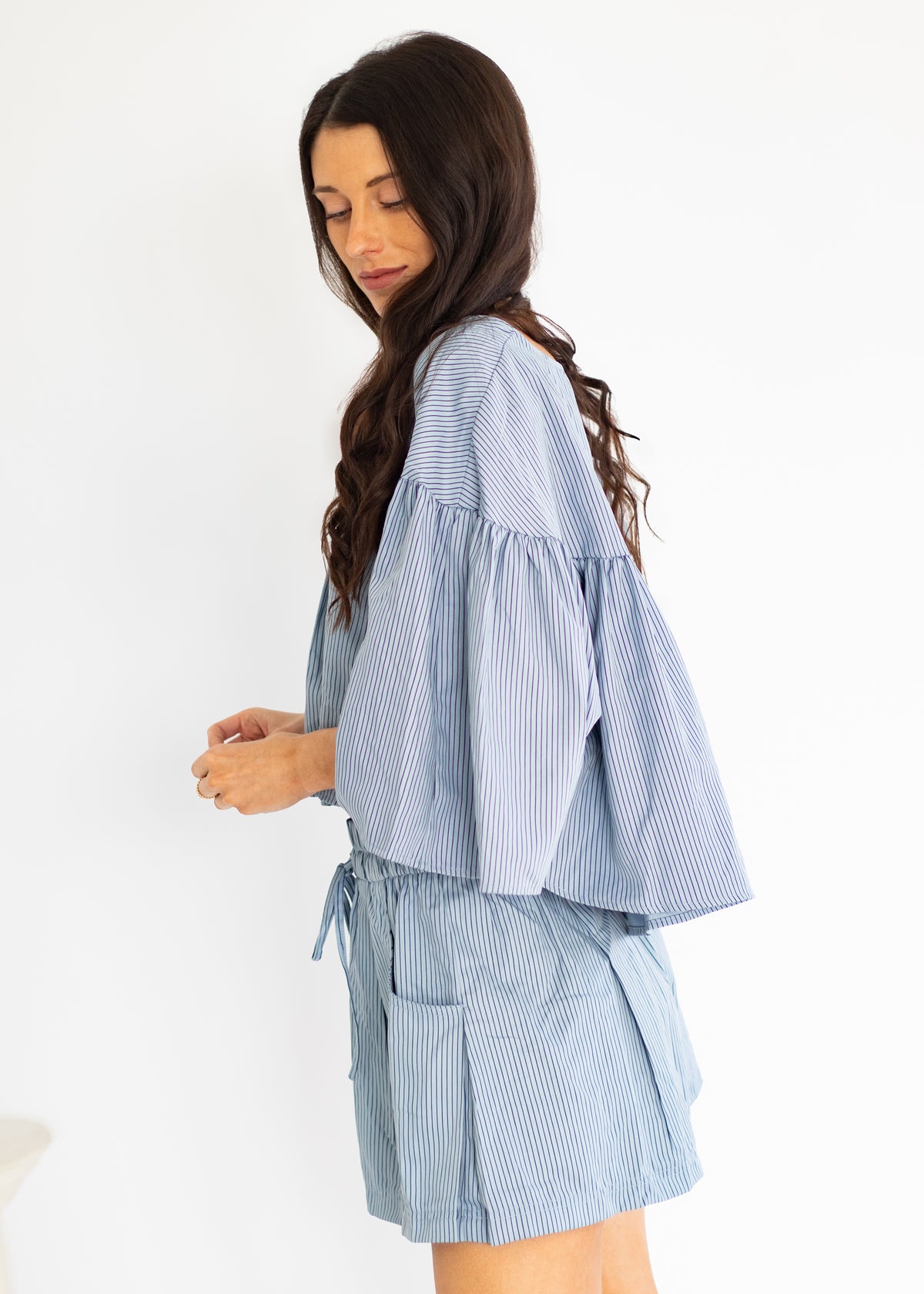 A woman wearing a light blue and white vertically striped matching set consisting of a short-sleeve blouse with dramatic ruffled sleeves and matching shorts, photographed against a plain white studio background.