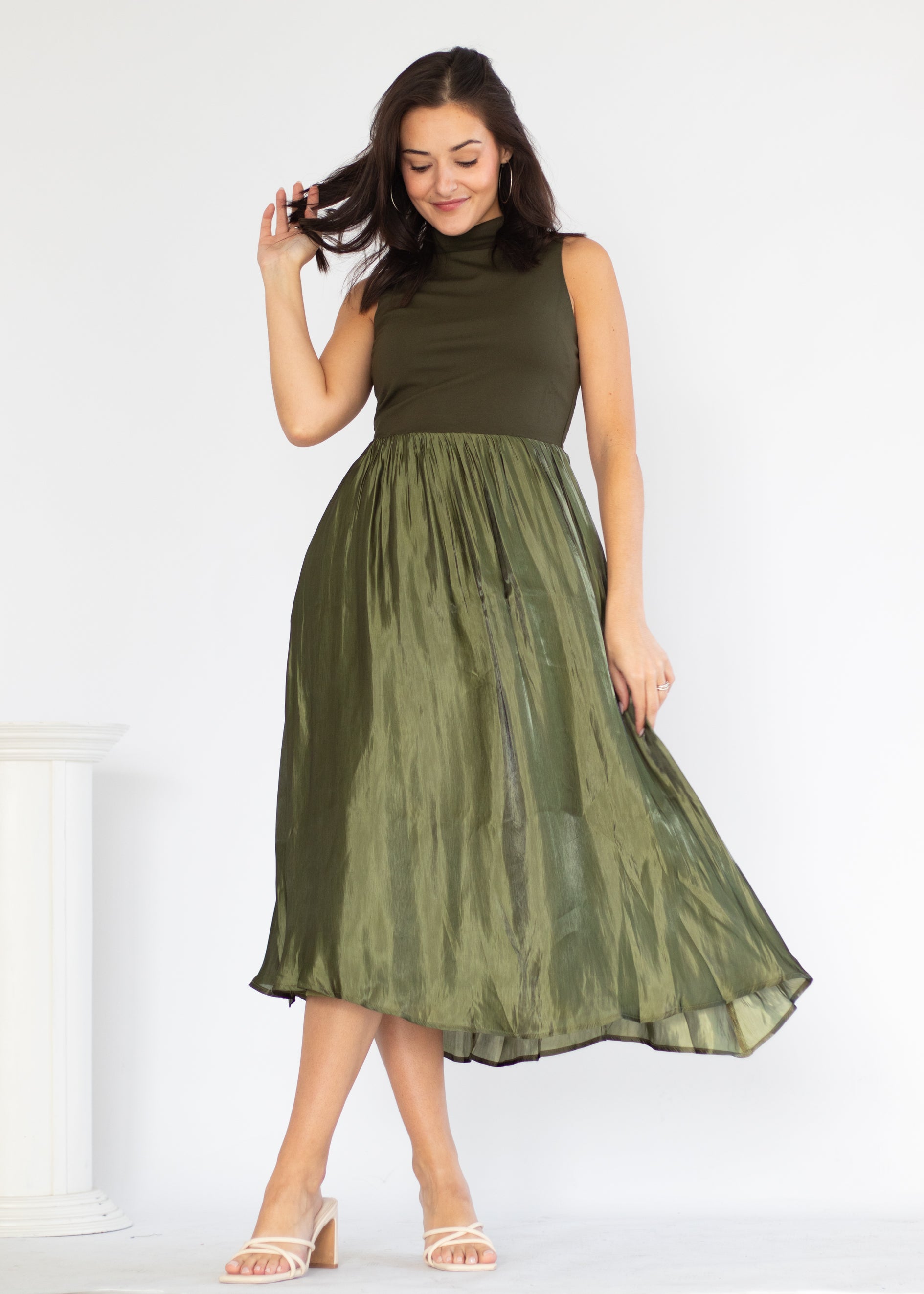 A woman modeling a sleeveless olive green midi dress with a high mock neck and a flowing, semi-sheer skirt, standing against a white studio background.