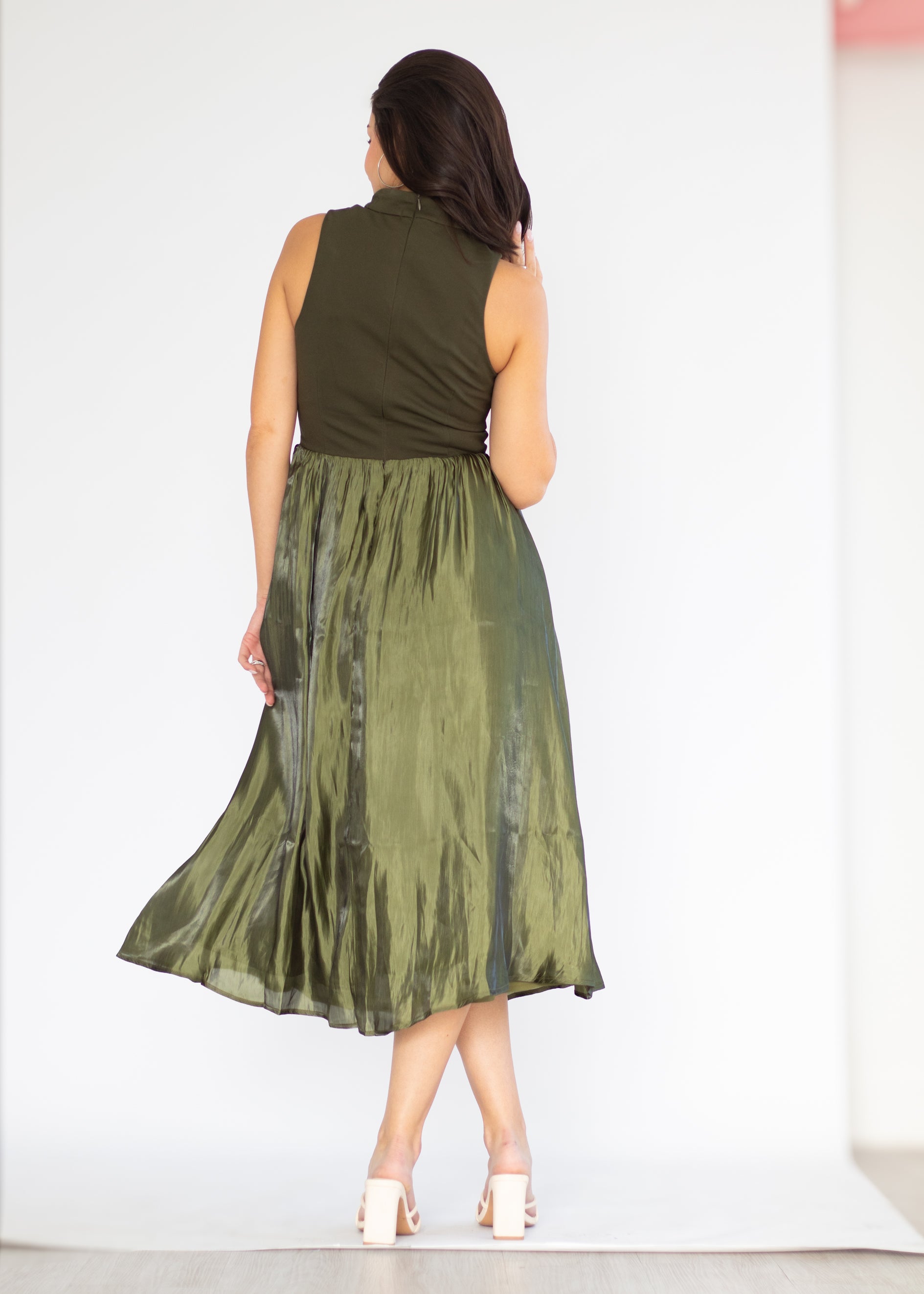 A woman shown from behind wearing a sleeveless olive green midi dress with a structured bodice and a flowing, semi-sheer skirt, paired with white high-heeled sandals, set against a plain white studio background.