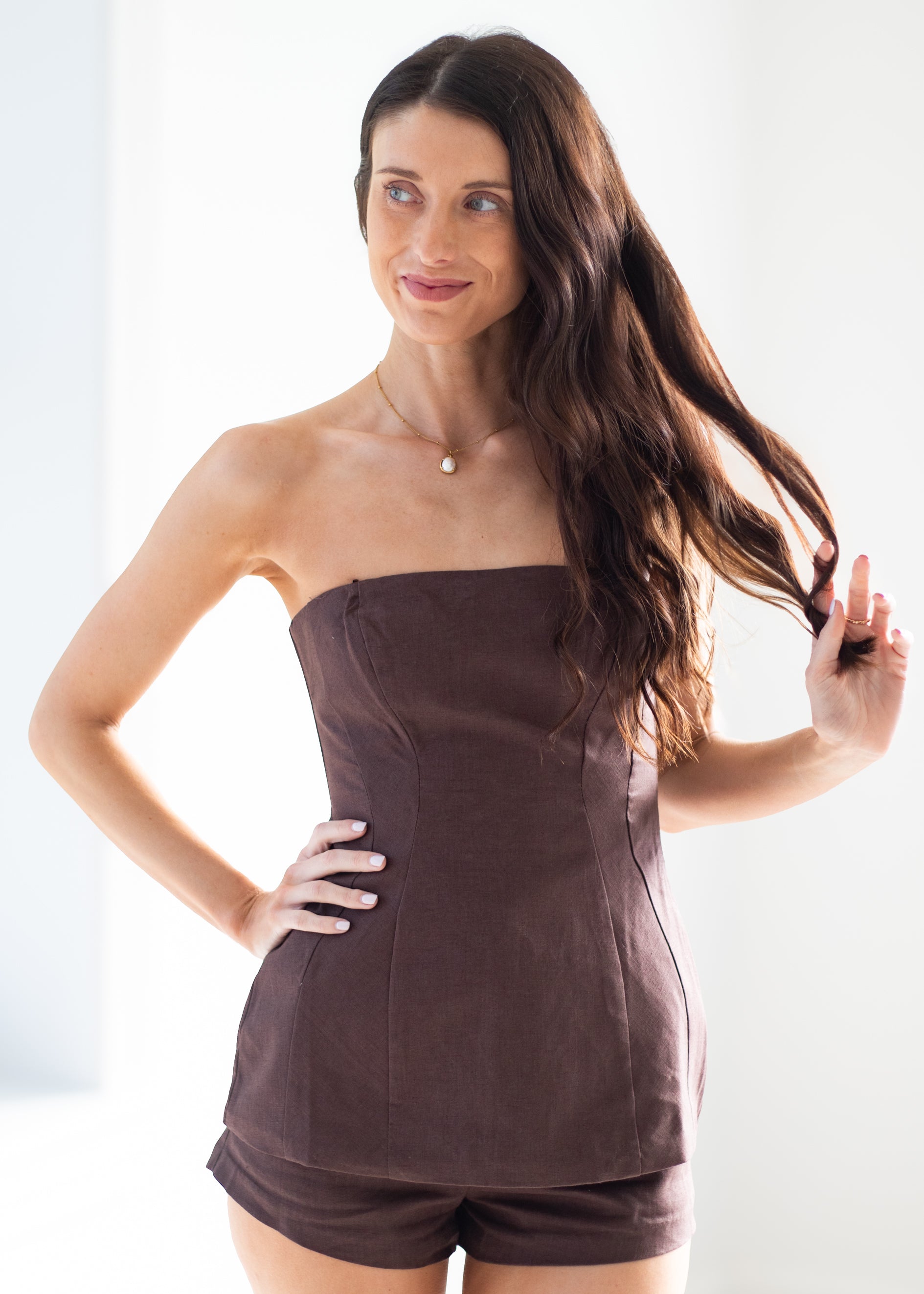 A woman modeling a dark brown strapless tube top and matching shorts set against a bright white background.